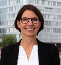 Porträt einer lächelnden Frau mit kurzer brauner Frisur und Brille, sie trägt ein weißes Oberteil und einen schwarzen Blazer vor modernem Gebäude.